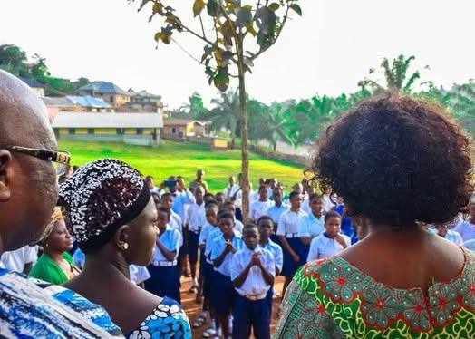 A speaker in traditional dress addressing rows of pupils in white and blue uniforms